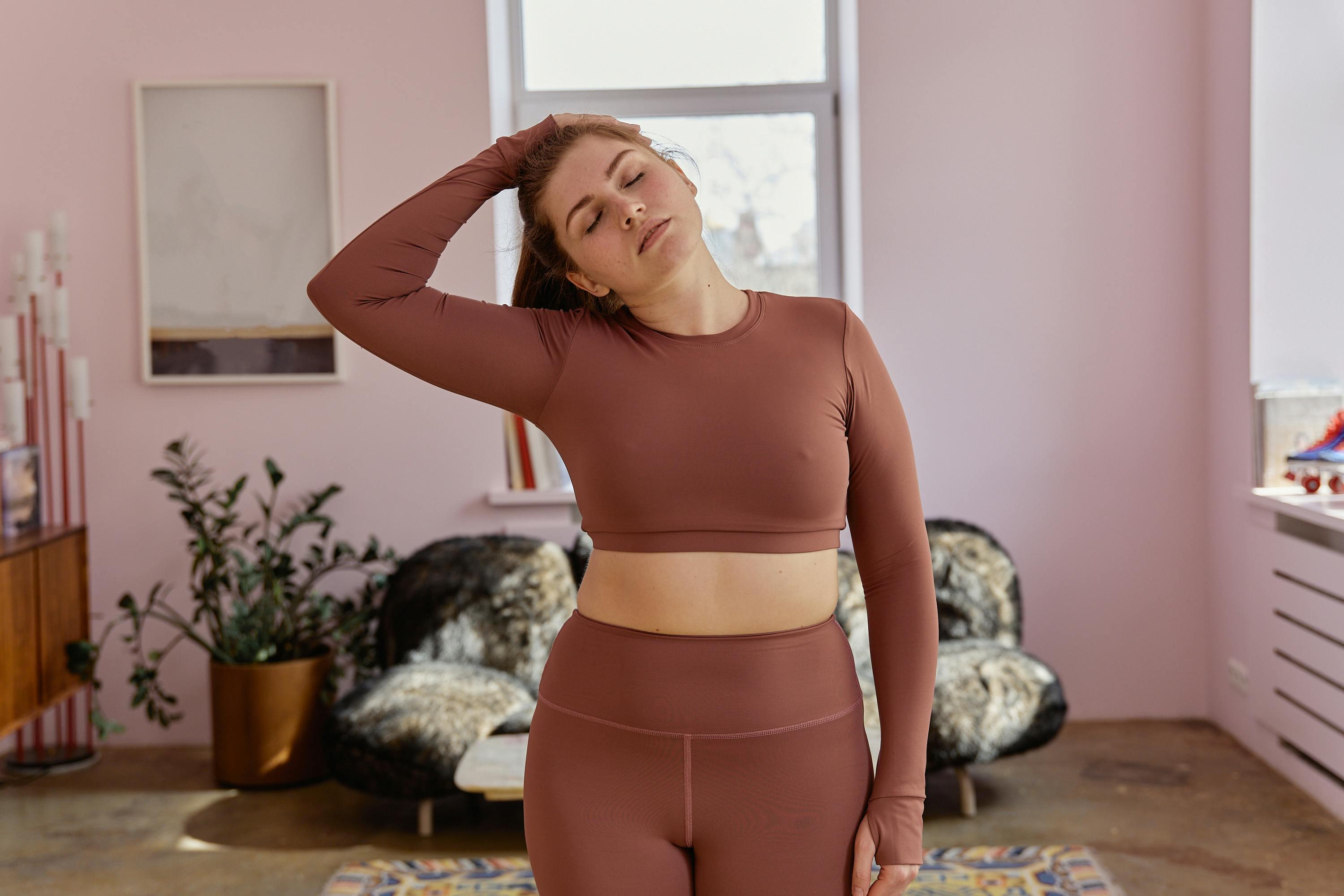 Mulher curvilínea usando conjunto fitness rosa empoeirado de mangas compridas realizando alongamento em sala de estar aconchegante.