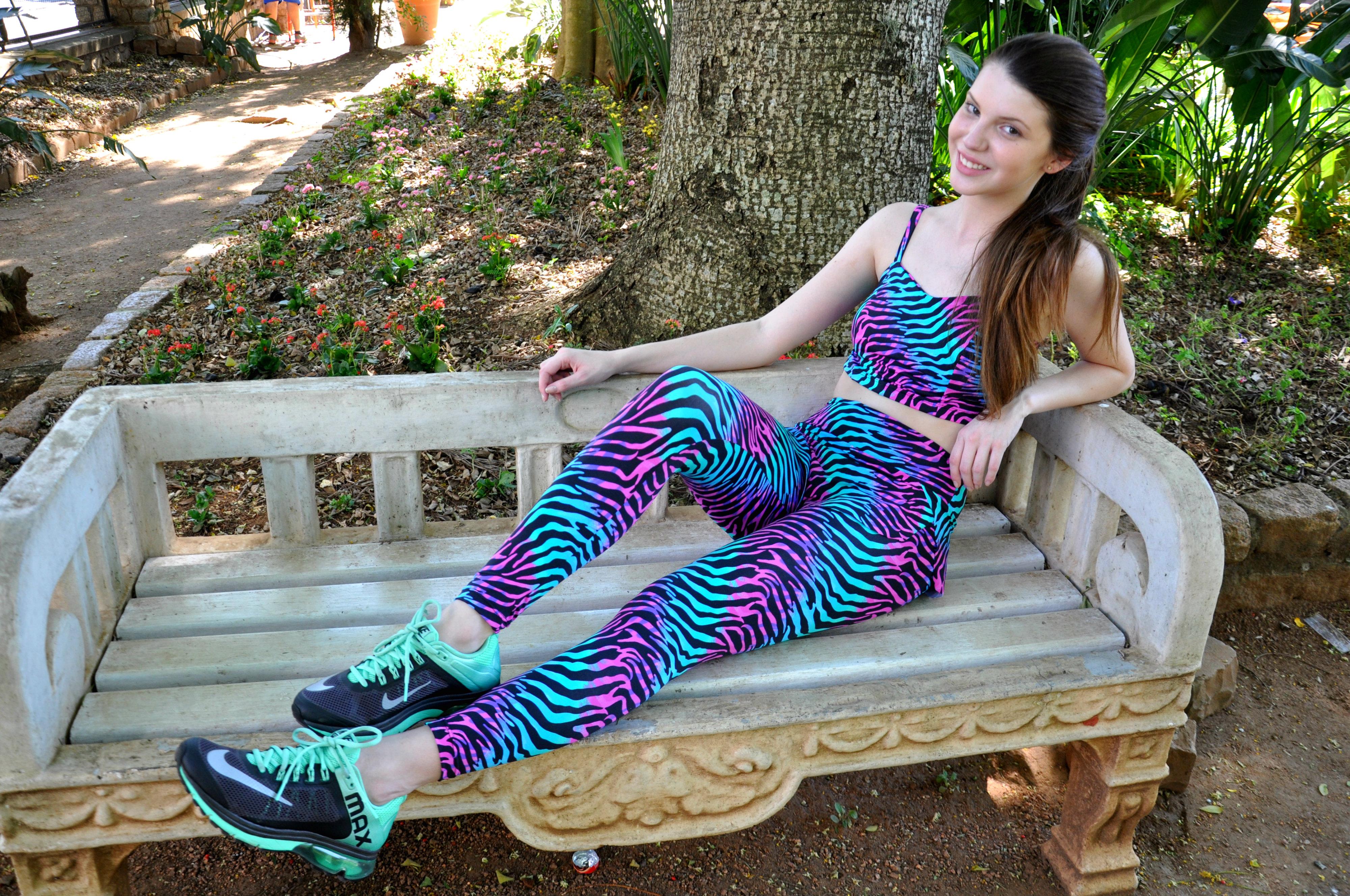 Jovem sentada em um banco de pedra ao ar livre, vestindo um top esportivo e leggings com estampa psicodélica vibrante de azul, rosa e verde.
